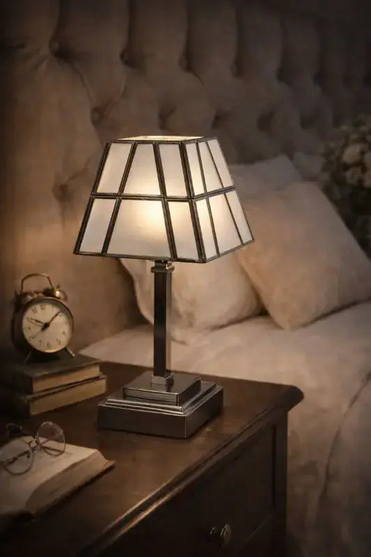 Table lamp with a decorative shade on a bedside table next to an alarm clock and books, with a bed in the background.